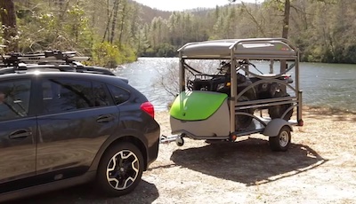 A SylvanSport GO hitched to a car with an ATV loaded on the gear deck with a lake in the background
