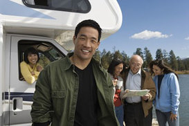 A man posed in the foreground, while his family is behind him (three members gathered together, looking at a map; and one sitting inside a white RV). A lake and trees are in the background.
