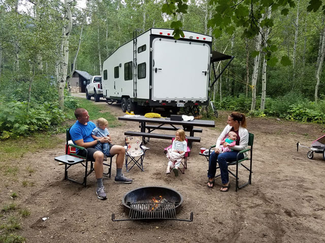 A family camping in the woods and sitting in front of a fire pit. A white camping trailer, attached to a truck, is behind them.