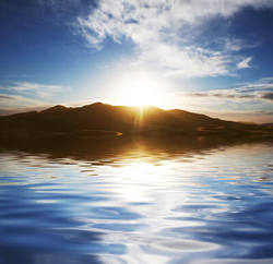 Image of a mountain looming over a lake, with a sun rising over it and casting a reflection in the water.