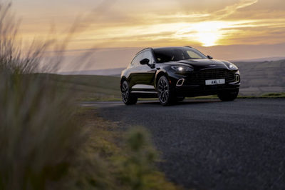 A black 2021 Aston Martin DBX traveling down a coastal road, with water and a sunset in the background.