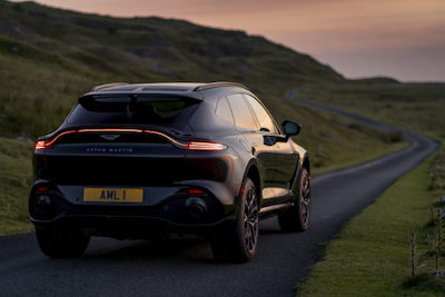 A black 2021 Aston Martin DBX driving down a coastal road at dusk, with green hills and water in the background.