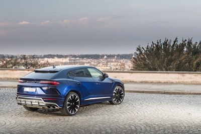 A blue 2021 Lamborghini Urus parked on a brick lot, with trees, shrubs, and sand positioned beyond.