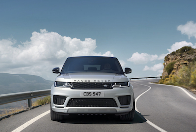 A grey 2021 Land Rover Range Rover Sport driving down a coastal road, with mountains and clouds in the background.