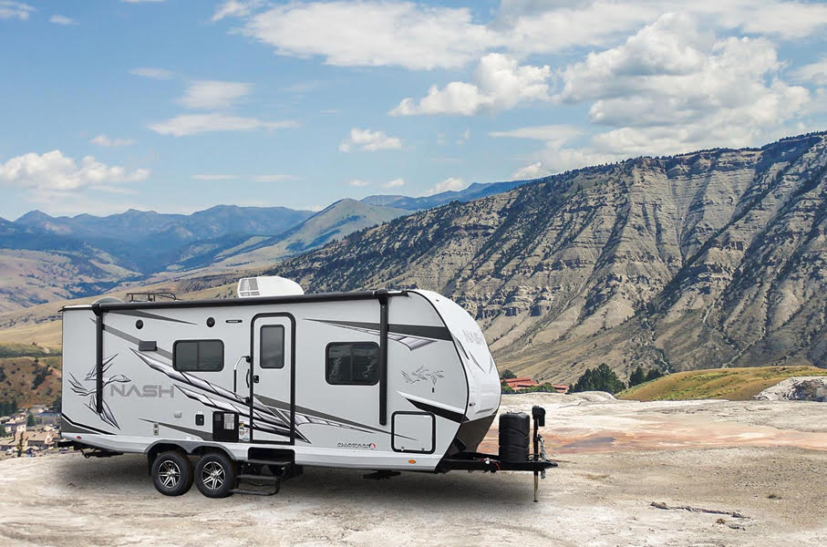 A white, grey, and black Nash travel trailer parked on a rocky cliff edge, with mountains and a town in the background.