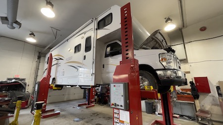 A white Ford-chassis motorhome raised on a column lift in the Skaha Ford repair facility. 