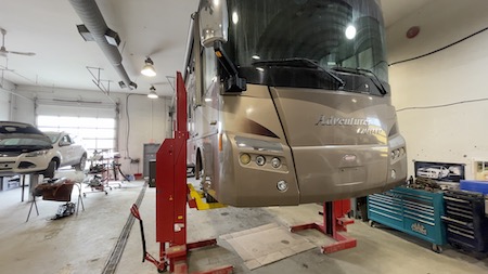 A brown Ford-chassis motorhome suspended on a column lift at the Skaha Ford repair facility.