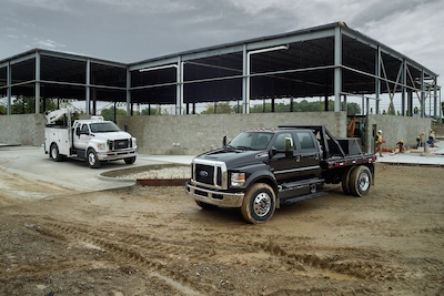 Two 2022 Ford F-650 trucks (one white and one black) parked at a work-site, with a building frame and construction workers in the background.