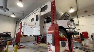 A white Ford-chassis motorhome suspended on a column lift at the Skaha Ford repair facility.