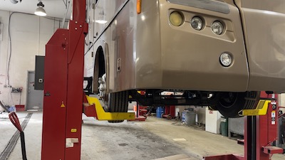 Close-up of a brown Ford-chassis RV being suspended on a column lift at the Skaha Ford repair facility, with its headlamps and front tire showcased.
