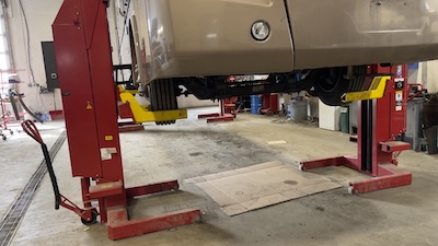 Close-up view of a Ford-chassis motorhome suspended on a column lift at the Skaha Ford repair center, with the front headlamp, tire, and bumper in view.