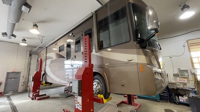 Side-view of a brown Ford-chassis motorhome suspended on a column lift at the Skaha Ford repair center.