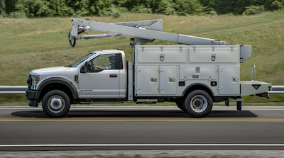 A white 2022 Ford F-550 XLT with a bucket attachment driving down a road, with grass and trees in the background.